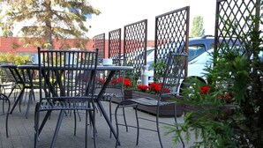 Espace extérieur aménagé avec des chaises et des tables en métal, entouré d’une clôture en treillis, avec une voiture et des fleurs rouges en arrière-plan.