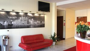 Un hall d’hôtel avec un canapé rouge, un comptoir de réception, un grand tableau représentant un paysage urbain en noir et blanc, une plante en pot et une poubelle.
