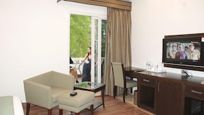 A hotel room with a sofa, armchair, dining table, TV, and a view of the outdoors. - Jht Hotels (New Delhi)