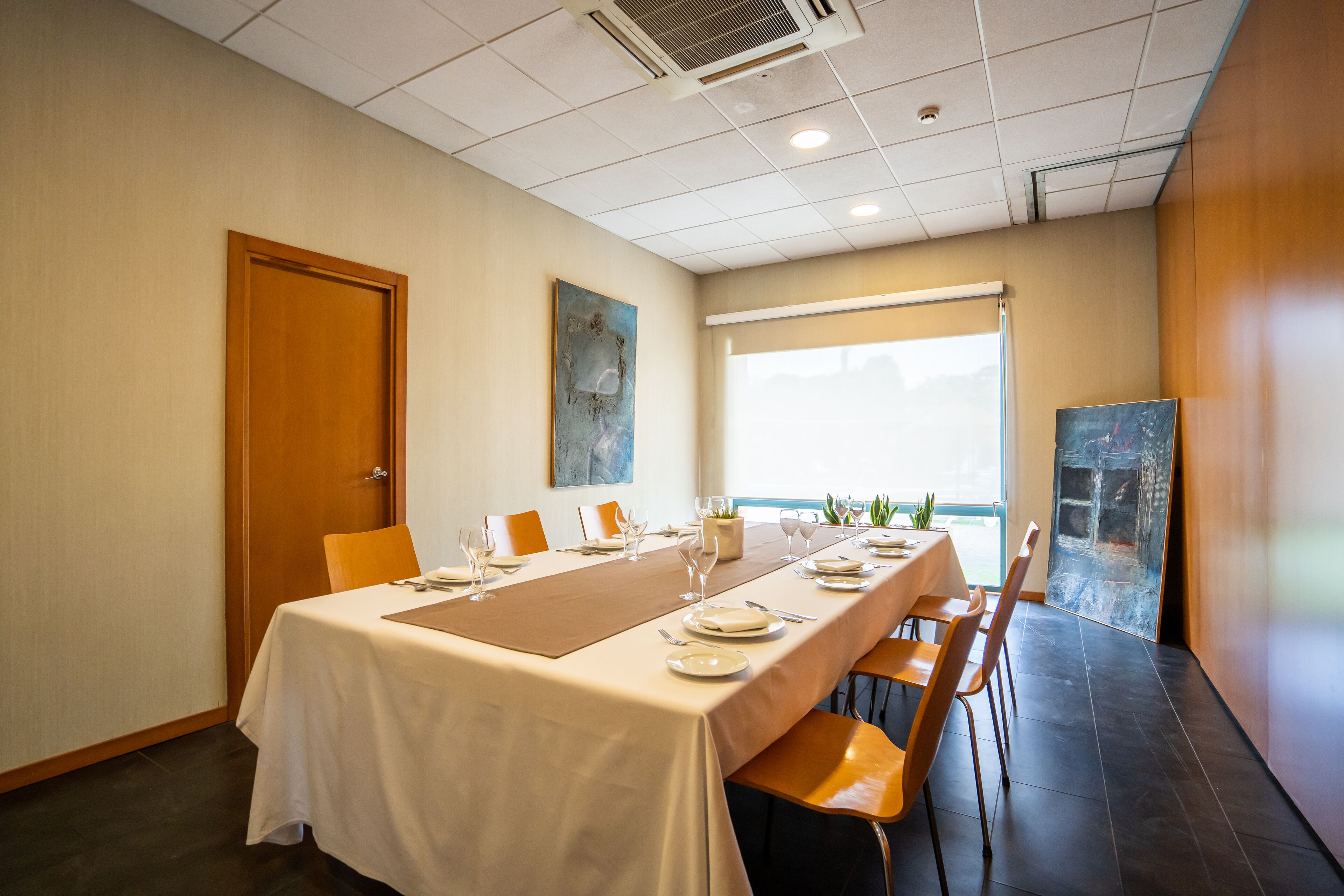 A conference room with a long table set for a meeting, equipped with chairs, a projector screen, and a large window providing natural light.