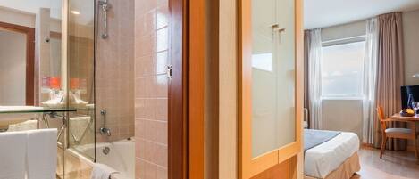 A hotel room with a glass shower enclosure, a bathtub, a bed with a wooden headboard, a dining table with chairs, and a large window with curtains.