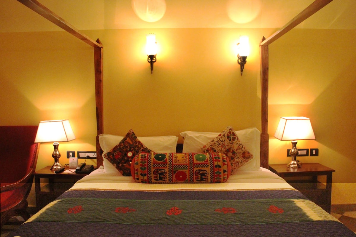 Habitación de hotel con cama con dosel, dos lámparas de noche, una silla y una mesita de noche con un teléfono.