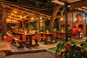 An outdoor dining area with wooden tables and benches, surrounded by greenery and a stone wall.