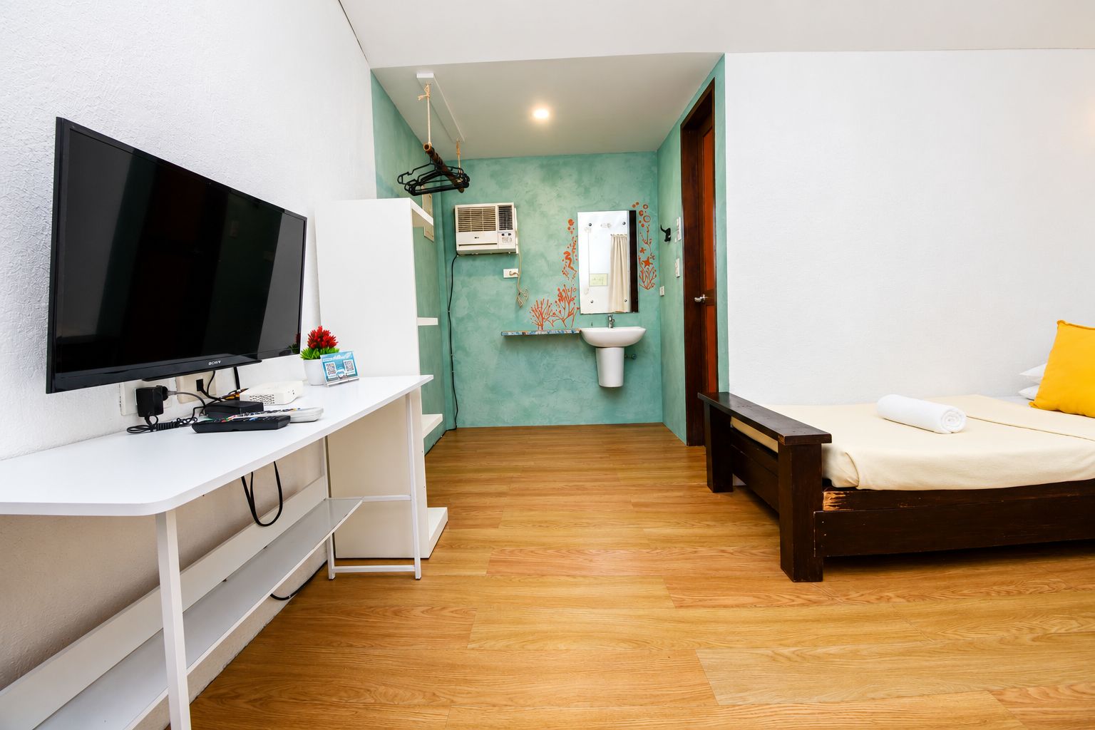 A bedroom with a bed, a desk with a TV, a bedside table, a bathroom with a sink and mirror, and a wooden floor.