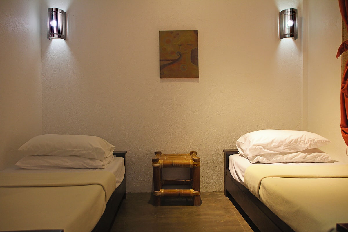 Habitación de hotel con dos camas individuales, un taburete de madera y dos lámparas fijadas a la pared.