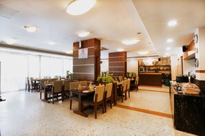 A spacious restaurant with multiple dining tables and chairs, a bar area, and a view of the city through large windows. - The HQ (Vasco da Gama)