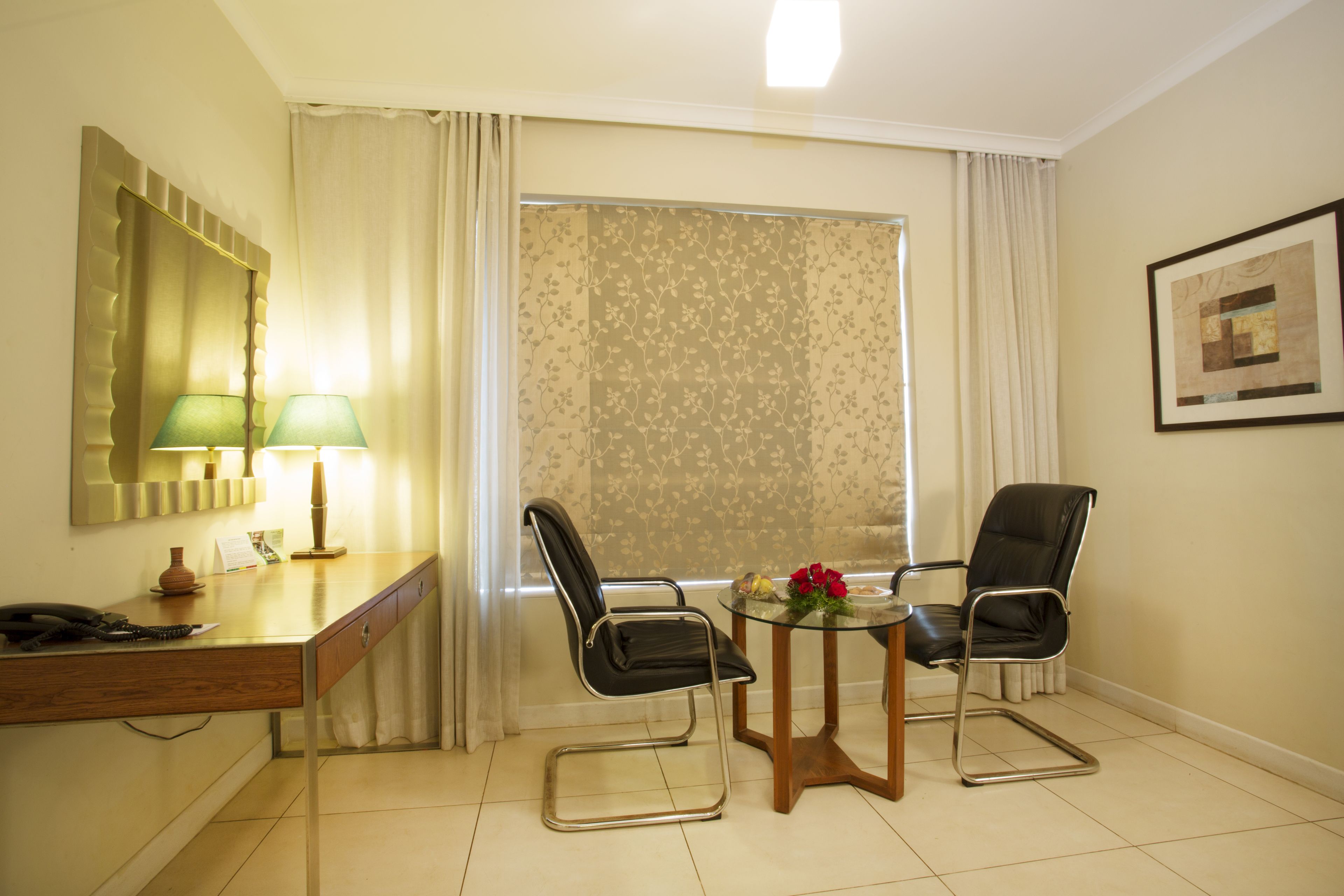A hotel room with a desk, two chairs, a glass table, a lamp, a telephone, and a window with curtains.
