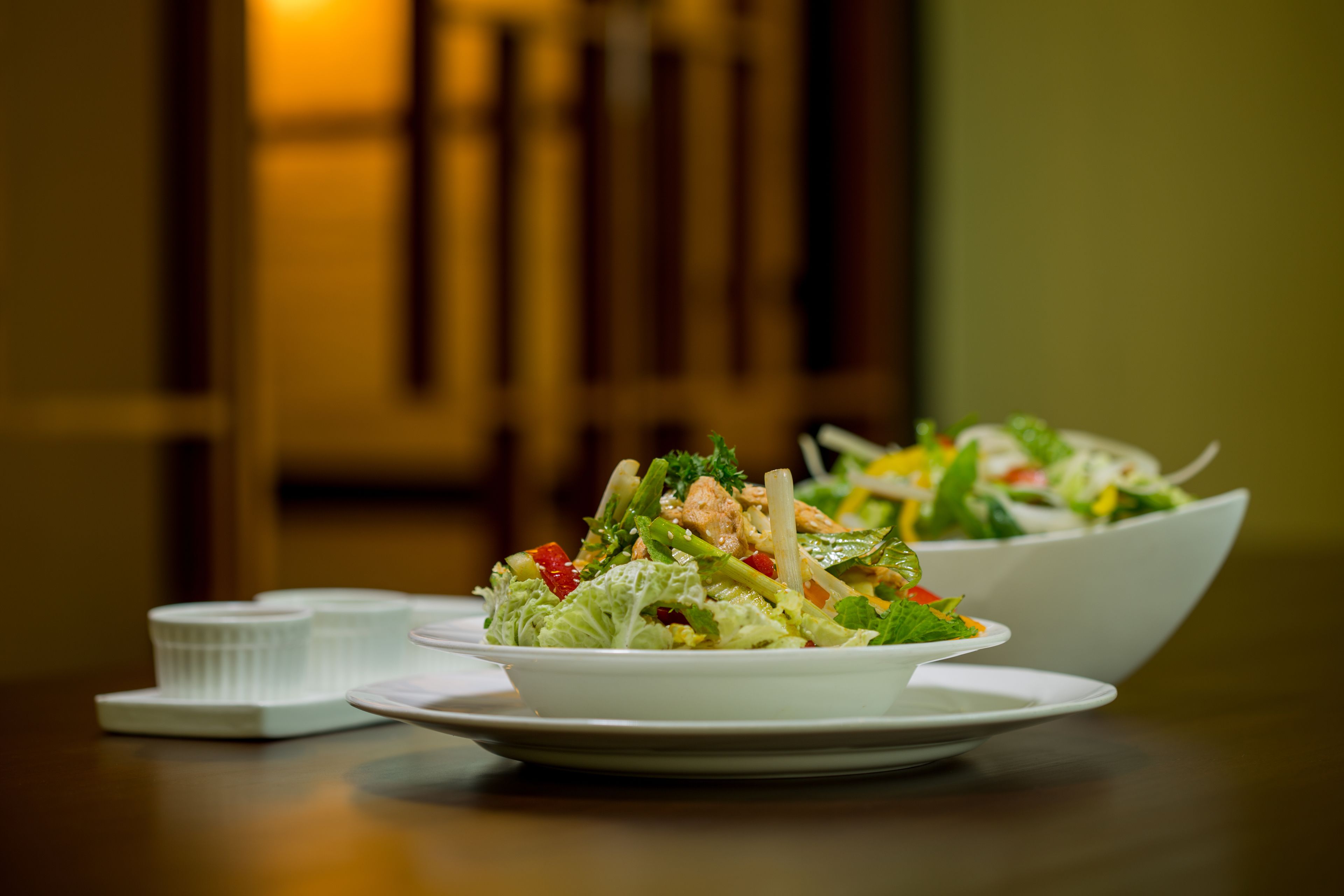 A dining table with two white plates of salad, a white bowl, and a white container.