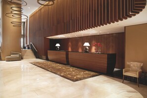 A modern reception area with a wooden reception desk, two lamps, a chair, and a staircase leading to upper floors.