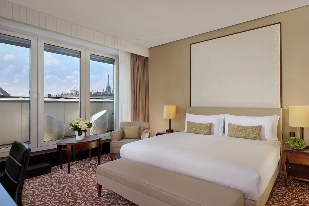 A hotel room with a large bed, a desk, a chair, and a view of a cityscape through large windows.