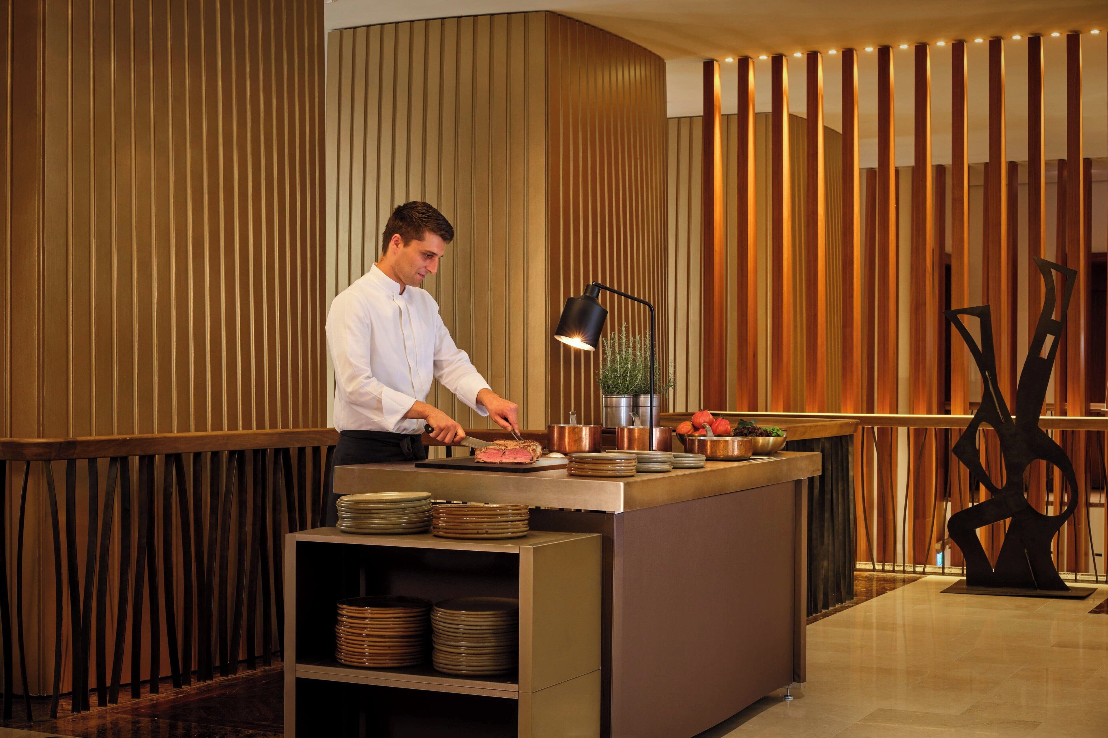 A chef preparing food at a counter with a modern design, including vertical wooden panels and a minimalist sculpture.