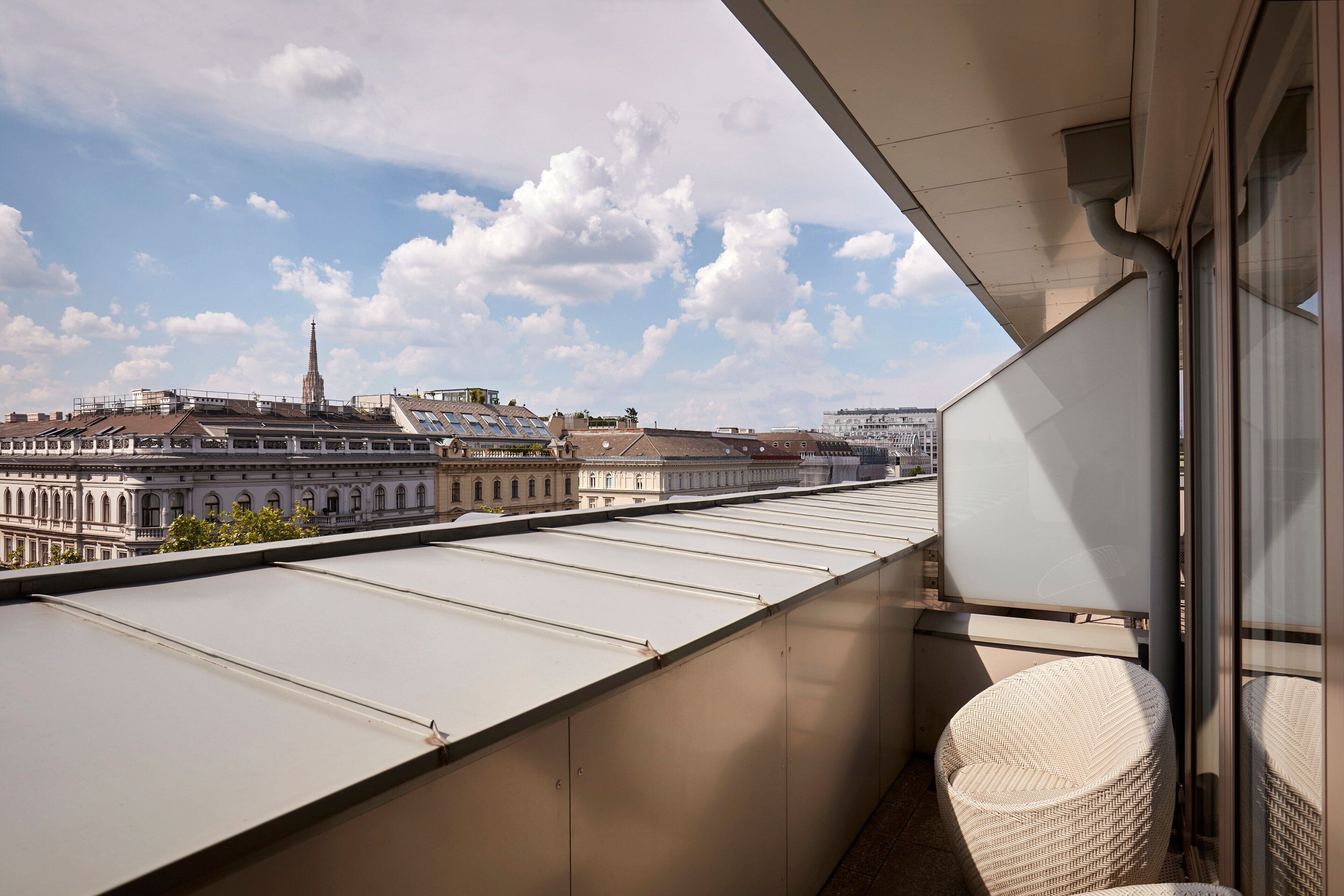 Un balcone con vista su un edificio storico con una guglia, cielo sereno e una sedia in vimini.