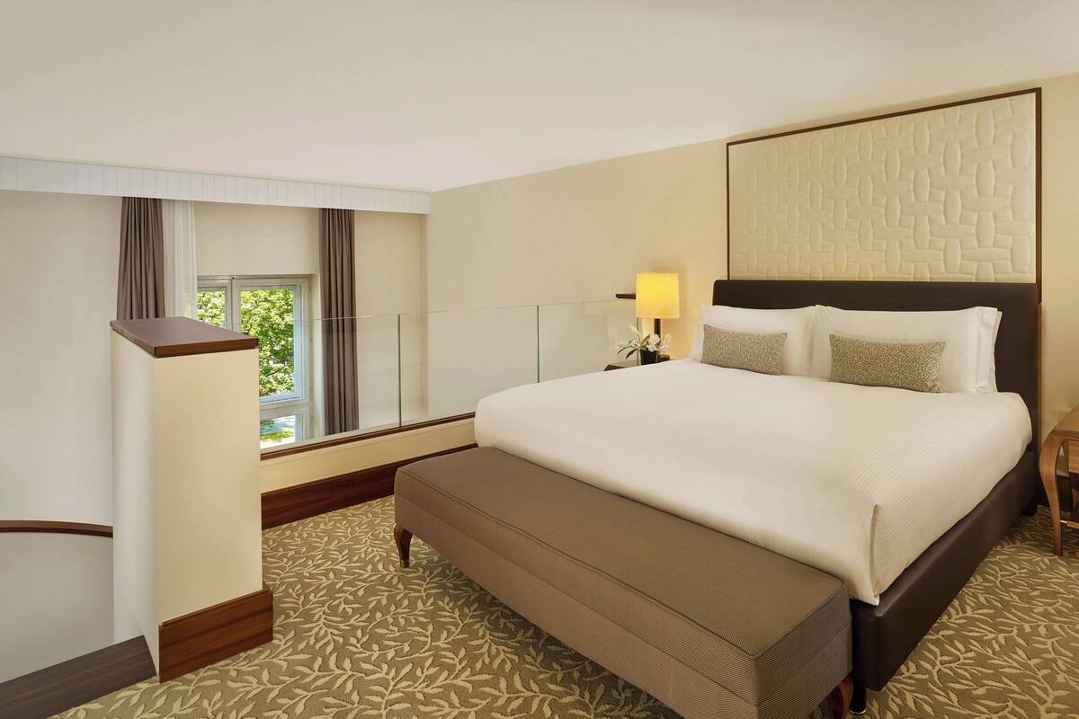 A hotel room with a large bed, a bench, a desk, a lamp, and a window with curtains.