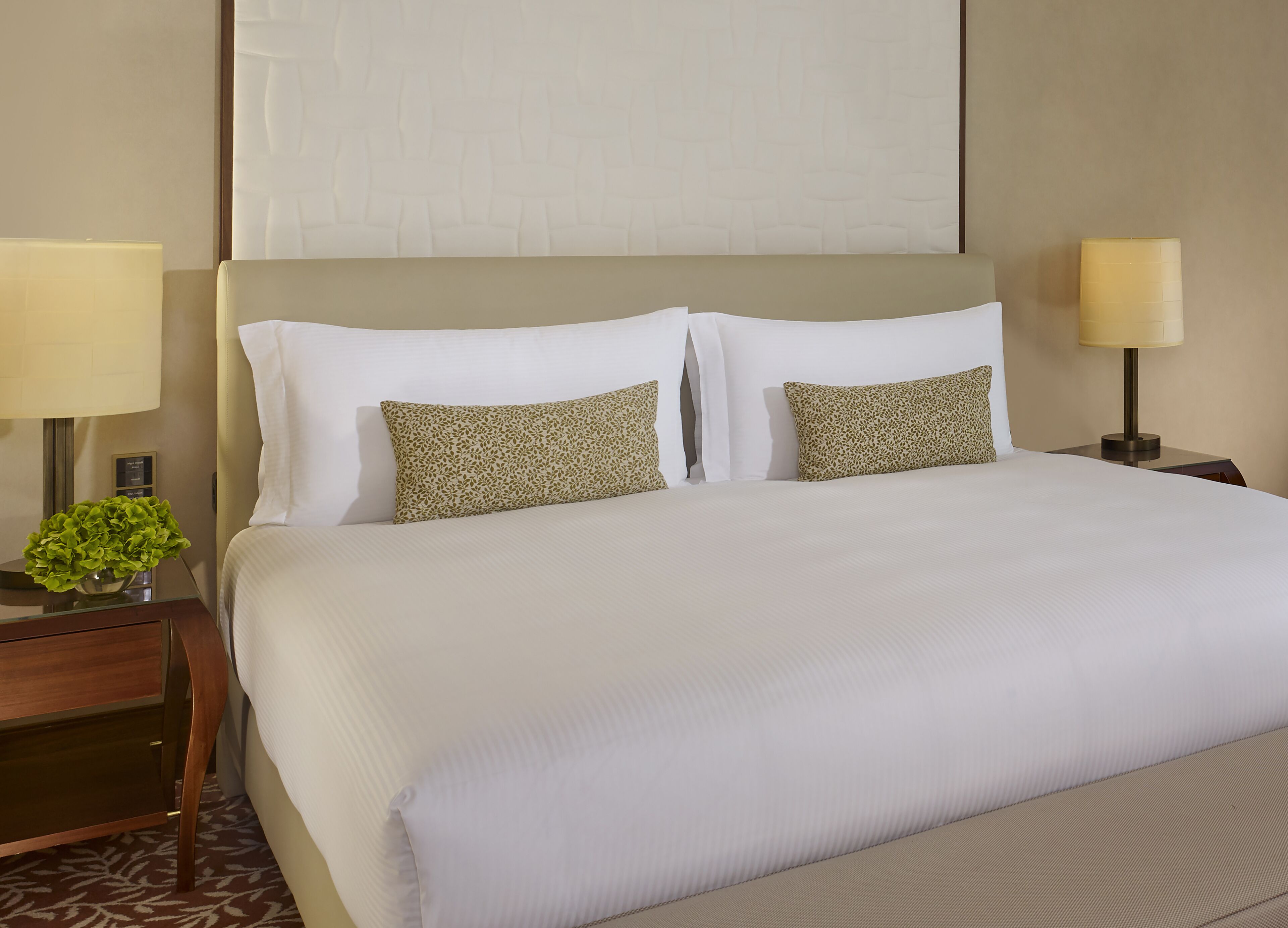 A hotel room with a large bed, two pillows, a nightstand with a lamp, and a small plant.