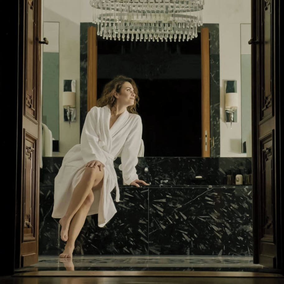 A woman in a white robe sitting on a black marble counter in a luxurious bathroom with a large mirror and a chandelier.
