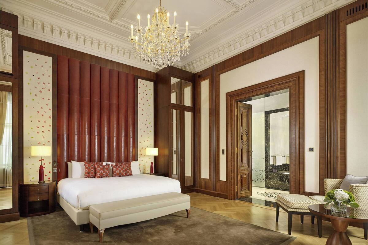 A spacious hotel room with a large bed, a chandelier, and a view of the lobby.