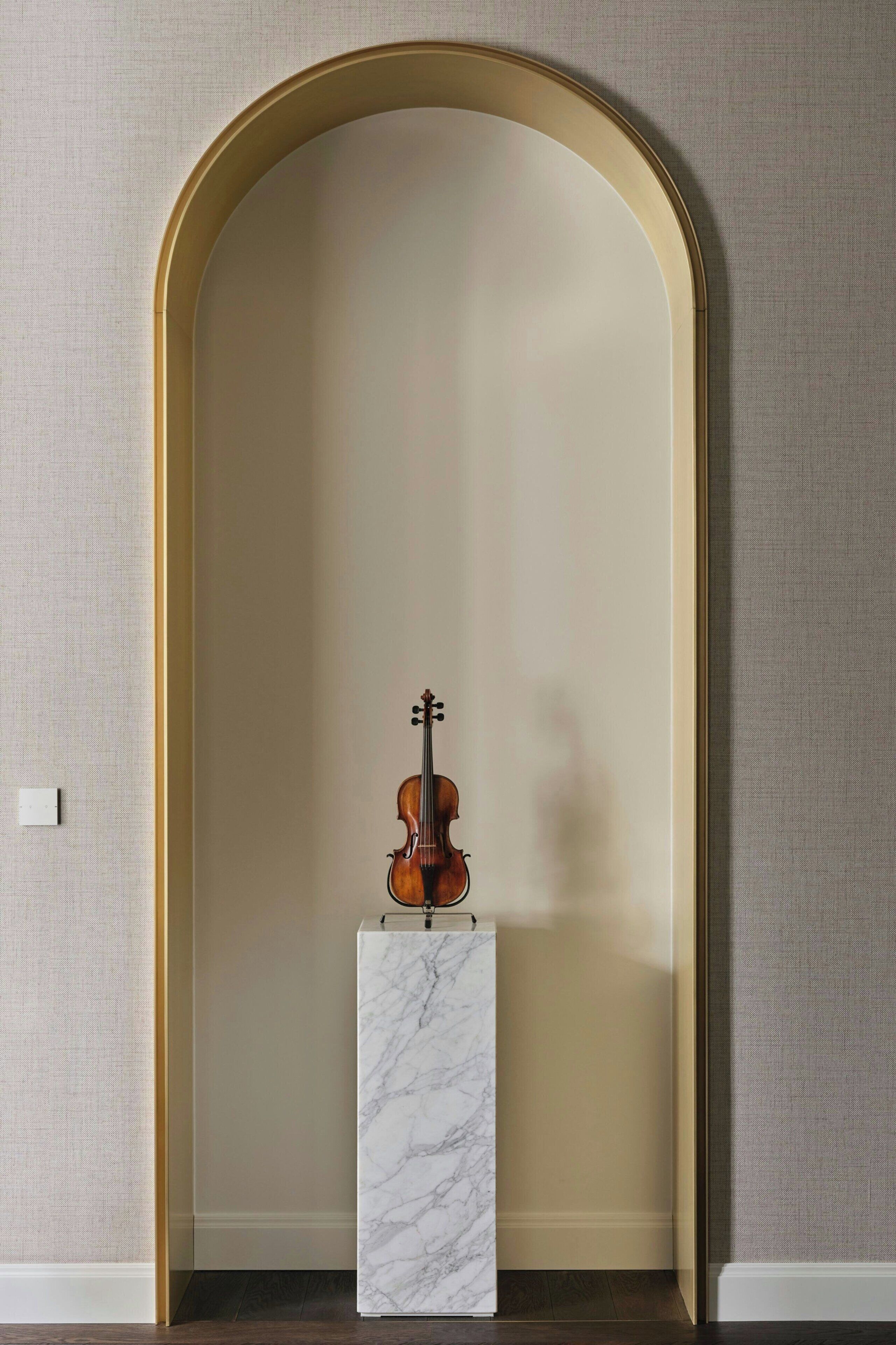 Un violín sobre un pedestal de mármol en una habitación con pared texturizada y suelo de madera.
