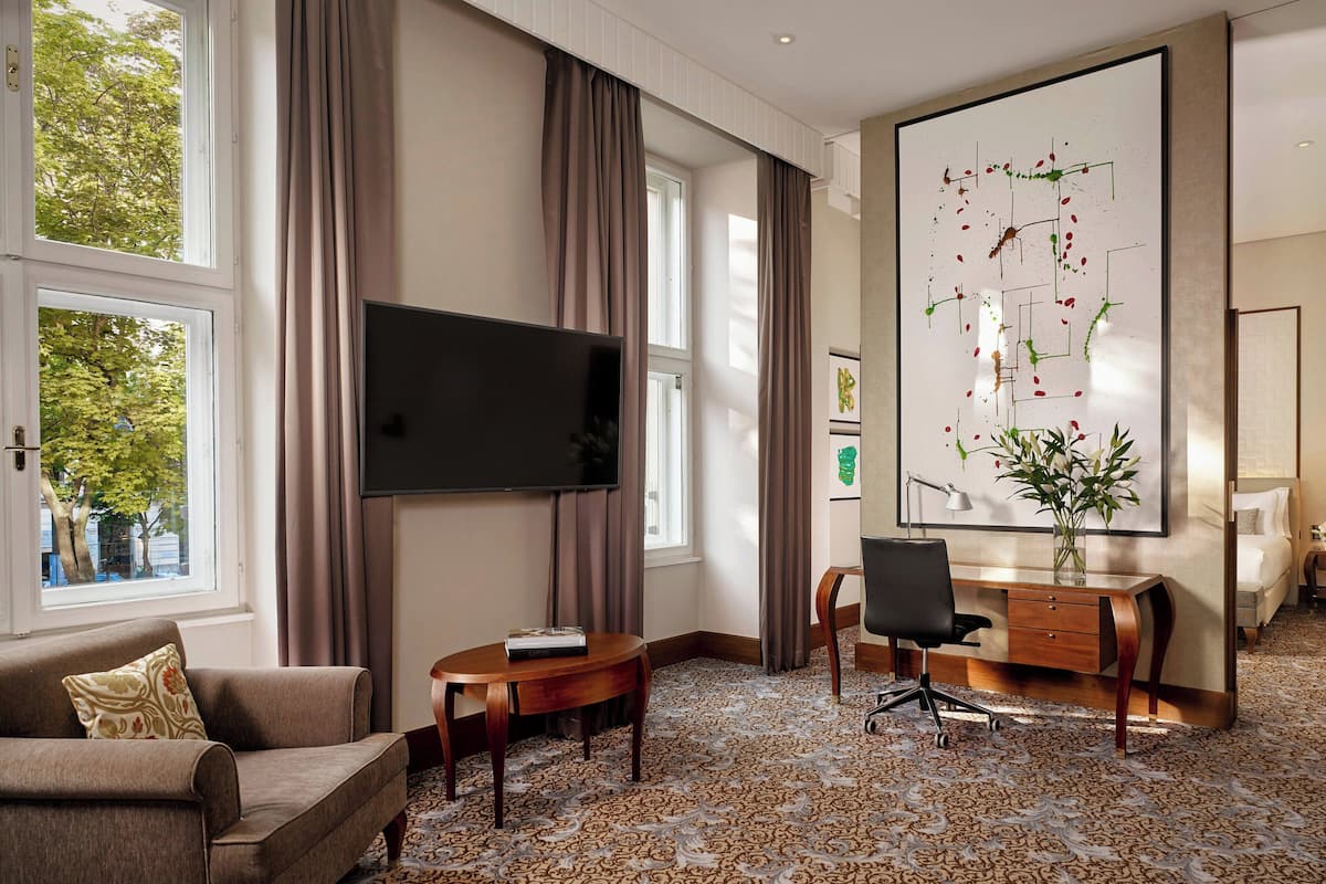 A hotel room with a large window, a flat-screen TV, a desk with a chair, a sofa, and a large abstract painting on the wall.