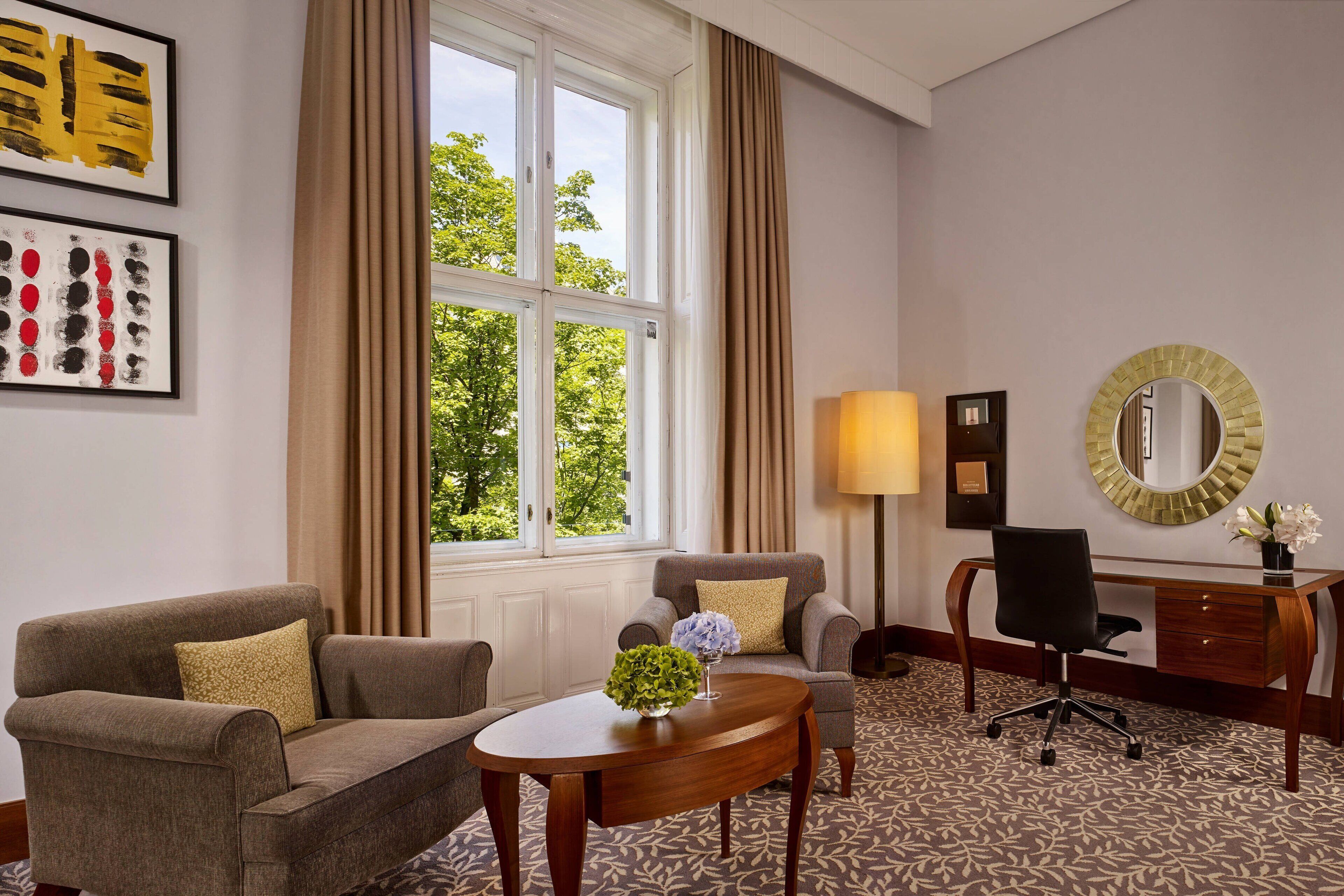 A hotel room with a sofa, a small table with a vase, a desk with a lamp, and a large window with curtains.