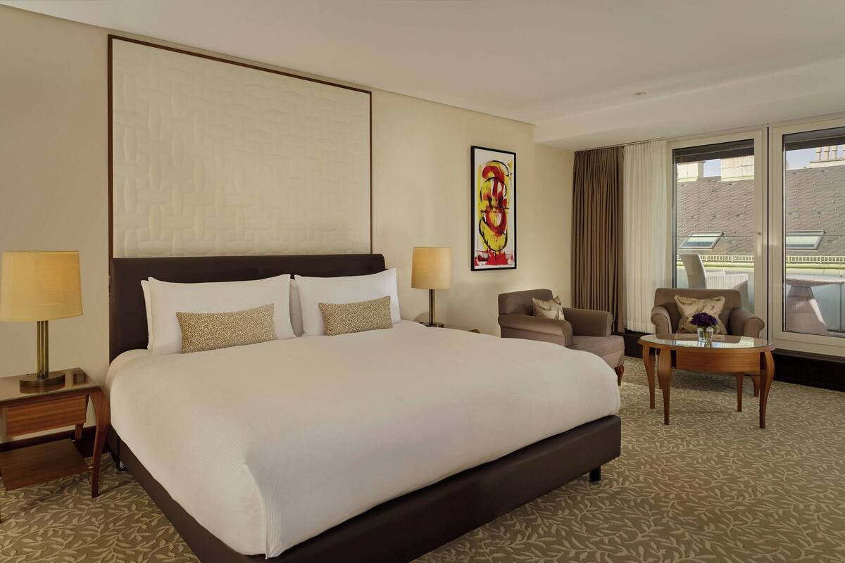 A hotel room with a large bed, two bedside tables, a chair, a small table, and a large window with curtains.