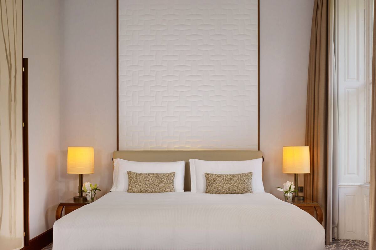 A neatly made bed with two pillows, a headboard with a textured pattern, two bedside lamps, and a nightstand with a vase of flowers.