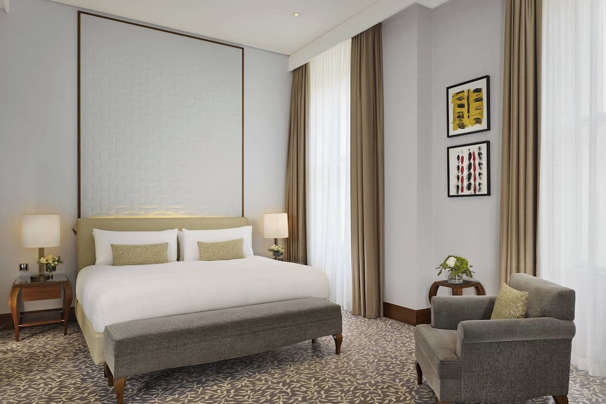A hotel room with a large bed, a grey armchair, a small side table, and two framed artworks on the wall.