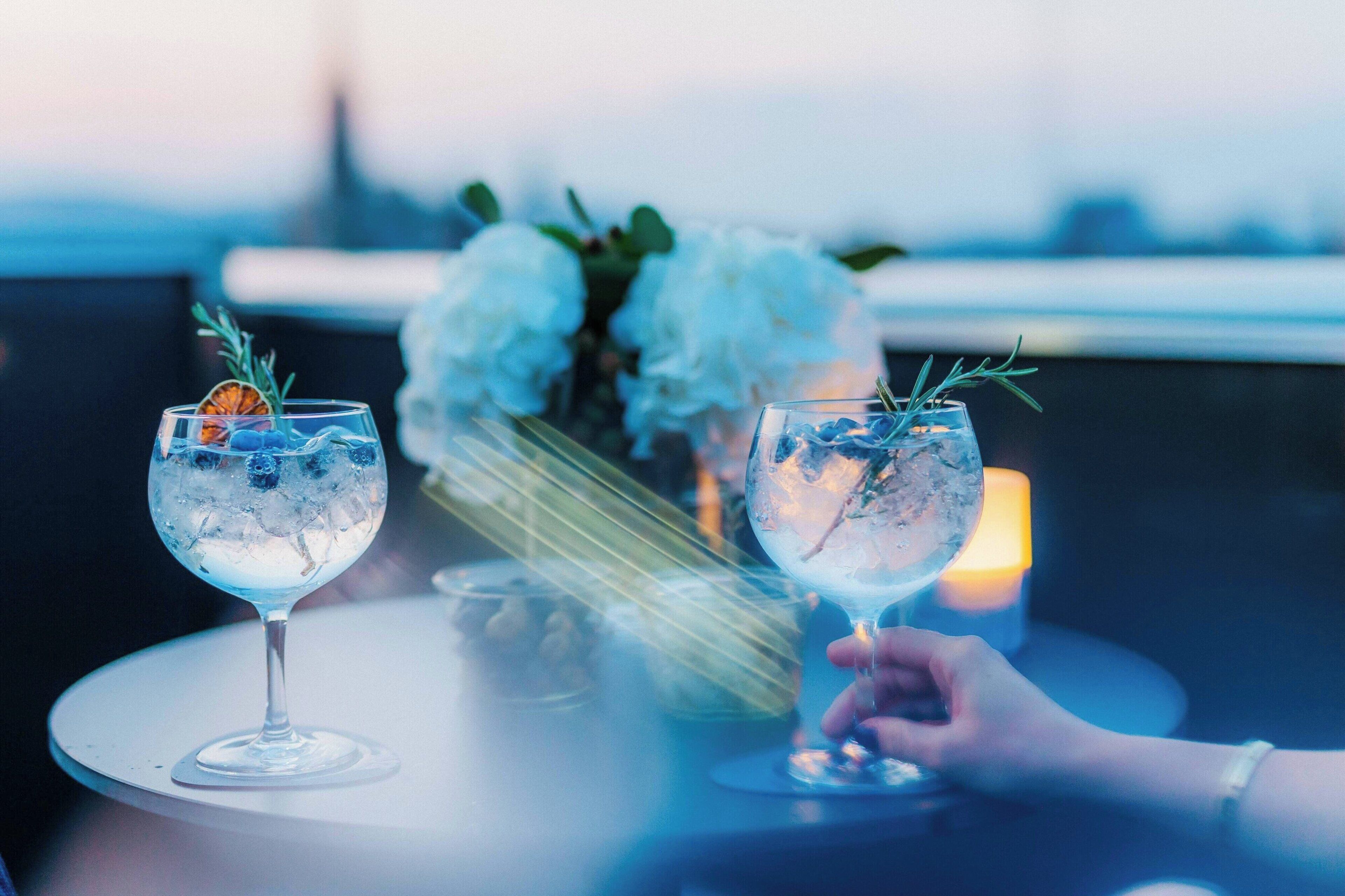 Two glasses of a clear beverage with ice and garnishes on a round table, with a blurred background of white flowers and a candle.
