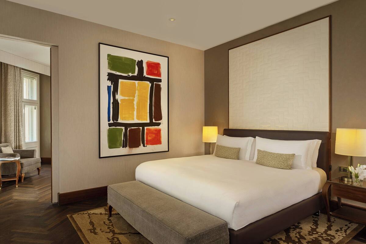 A hotel room with a large bed, two bedside lamps, a wall-mounted artwork, a bench, and a view of the hallway.