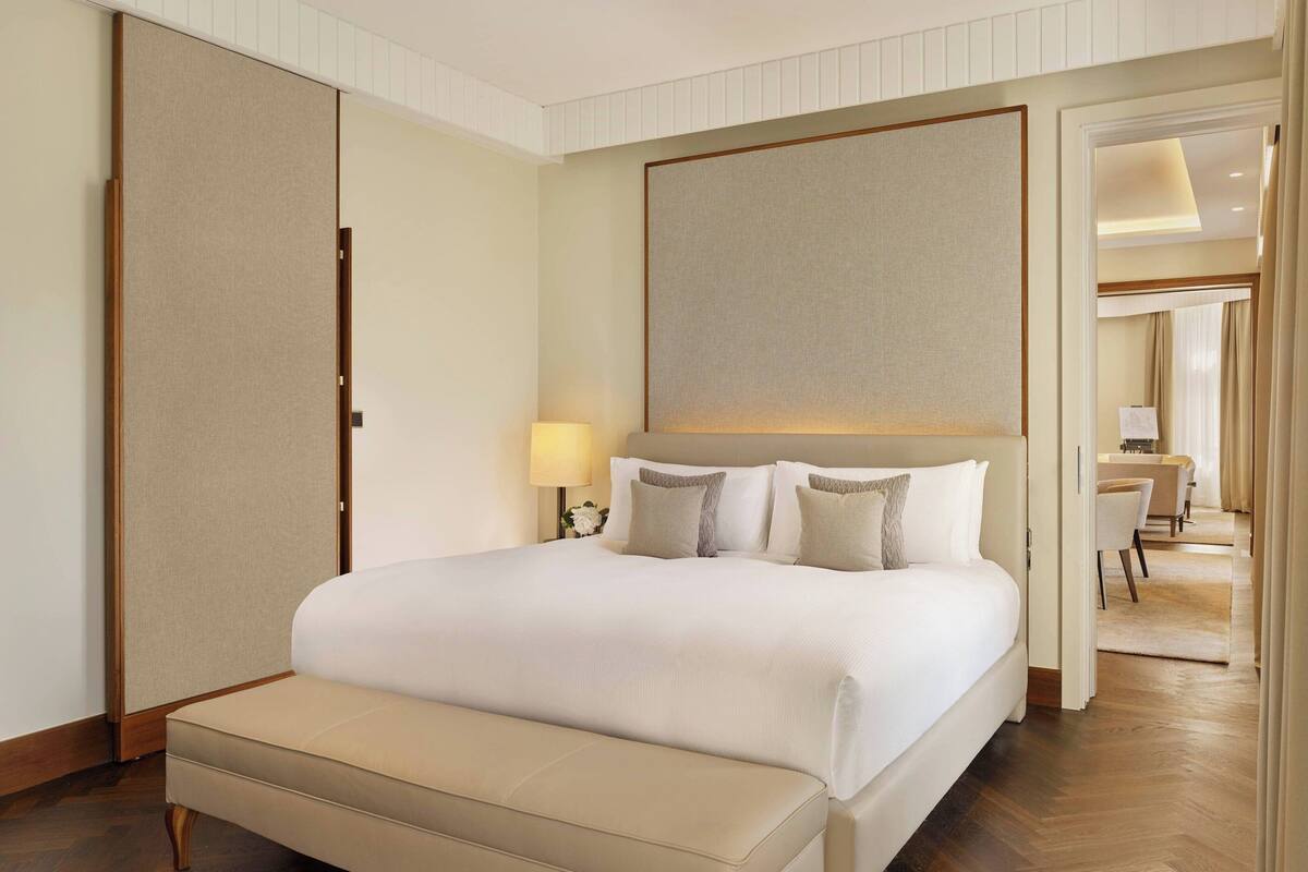 A hotel room with a large bed, a bedside table with a lamp, a bench at the foot of the bed, and a view into the adjacent room.