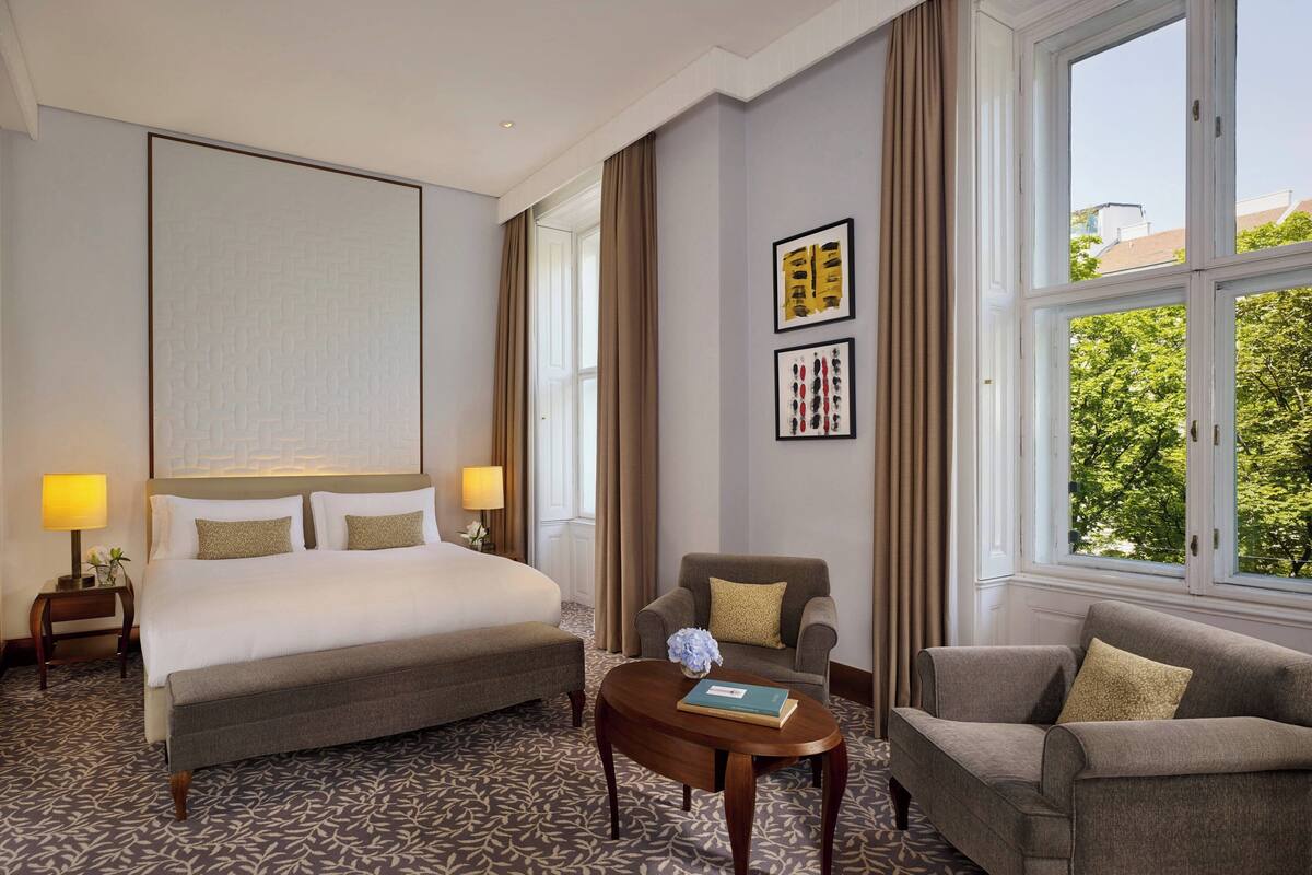 A hotel room with a large bed, two armchairs, a small table with a book, and a window with a view of trees.