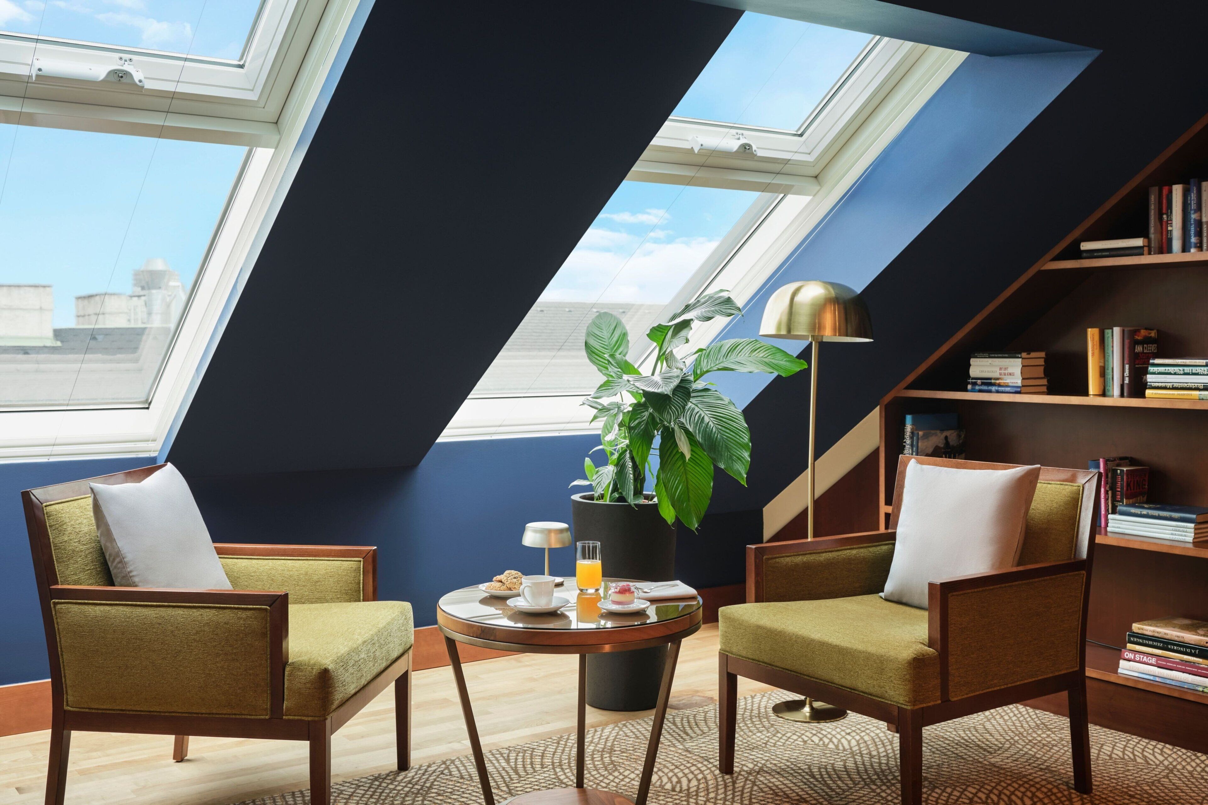 Um quarto com claraboia, duas poltronas, uma mesinha com uma planta, livros e uma luminária.