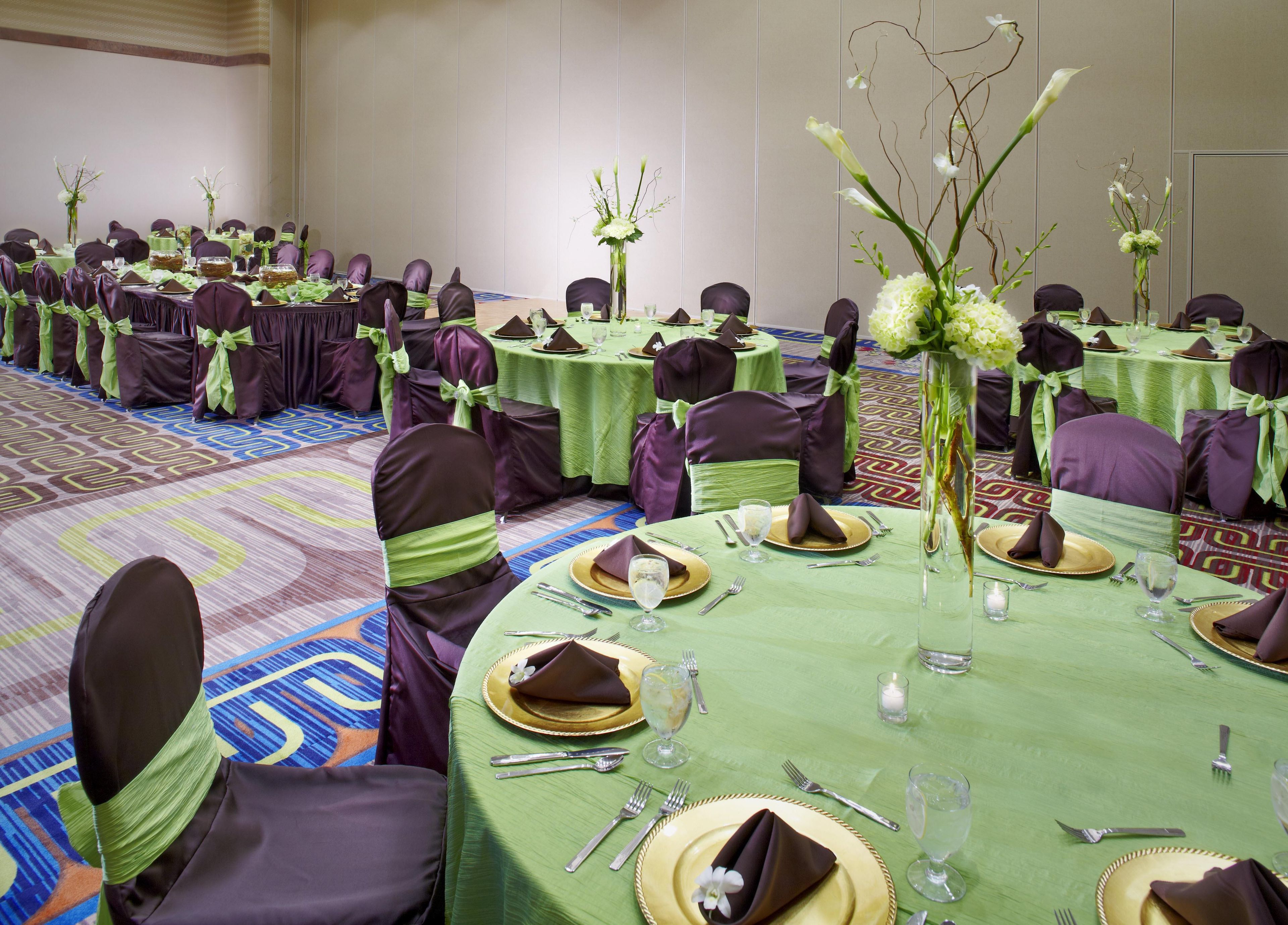 A banquet hall with round tables set for dining, each with green tablecloths, gold-rimmed plates, and purple chairs with green sashes.