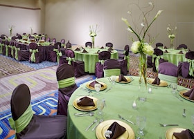 A banquet hall with round tables set for dining, each with green tablecloths, gold-rimmed plates, and purple chairs with green sashes.
