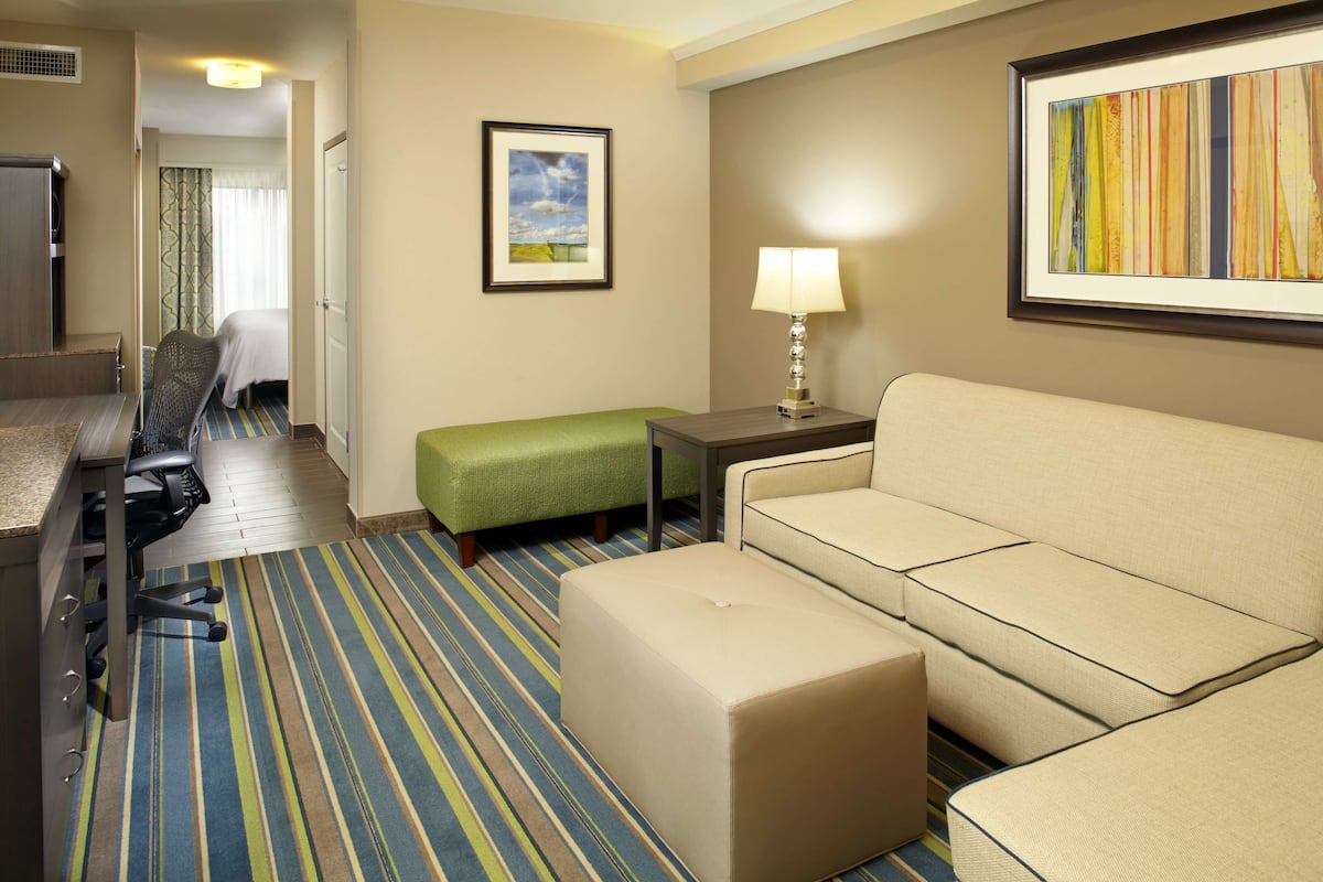 A hotel room with a bed, a green ottoman, a beige sofa, a desk with a chair, and a framed picture on the wall.