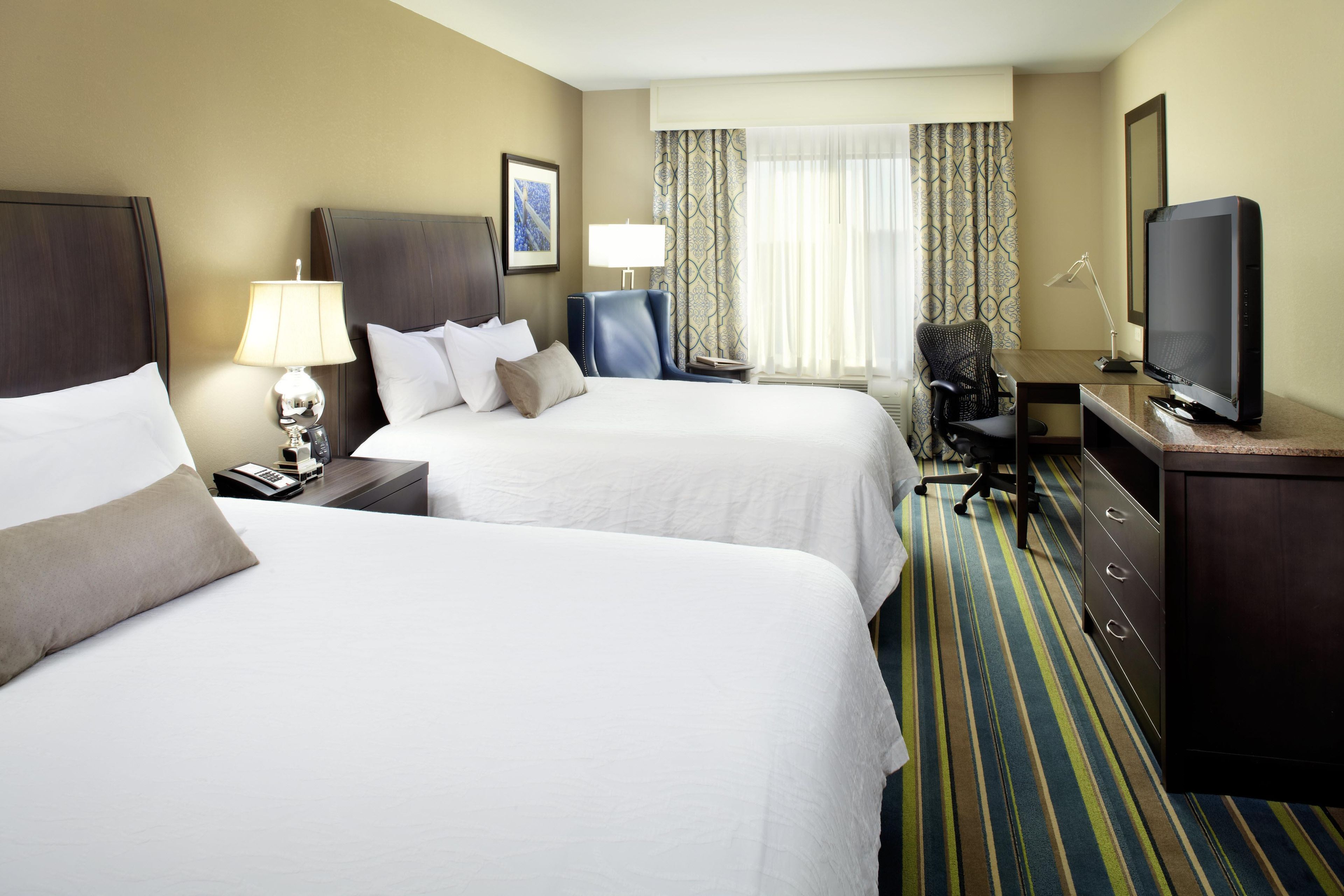 A hotel room with two beds, a desk with a television, a chair, and a desk lamp.