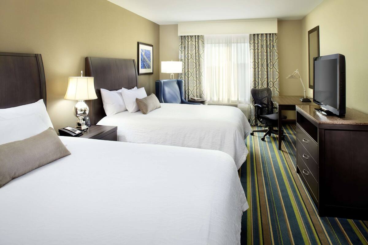 A hotel room with two beds, a desk with a television, a chair, and a lamp.