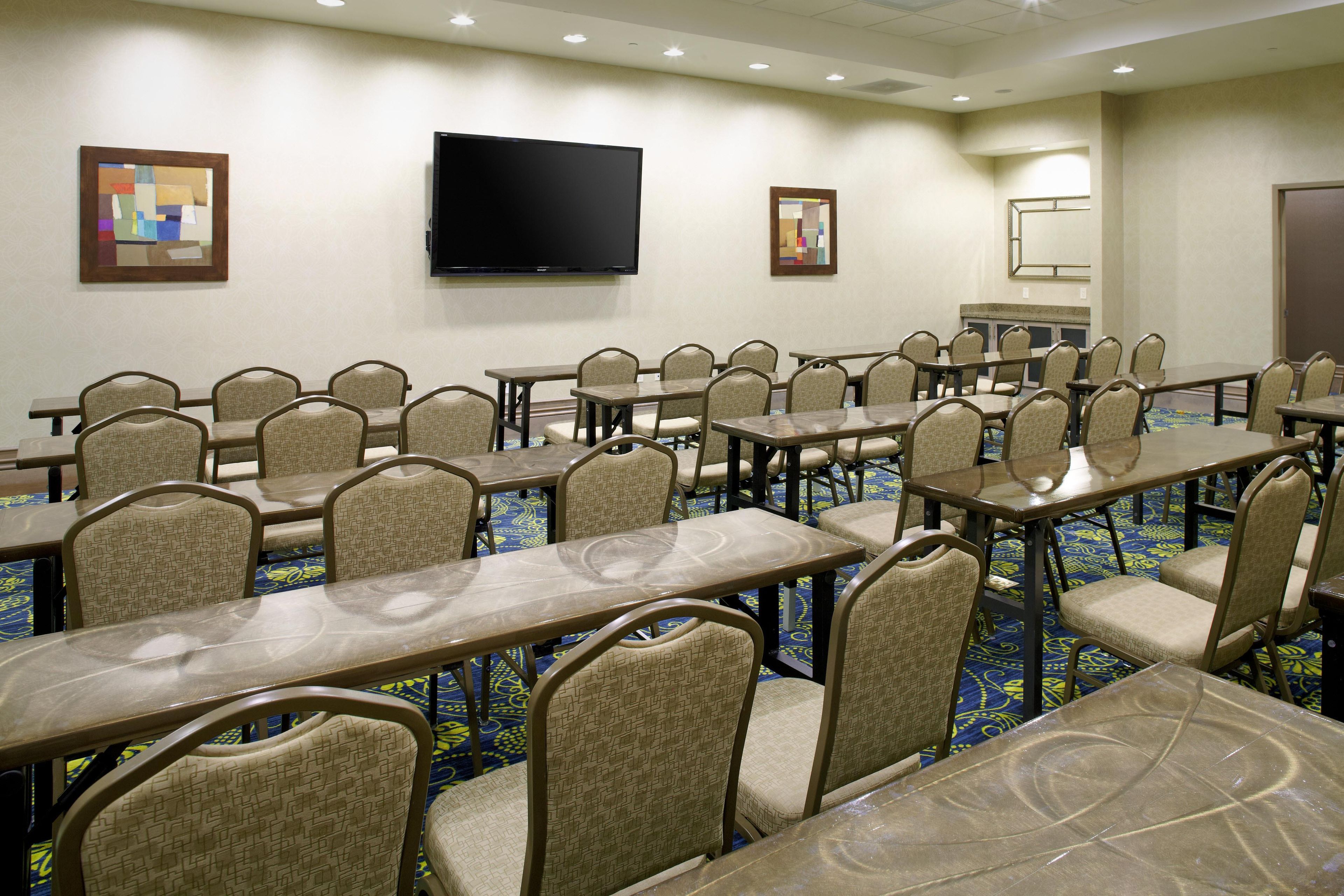 A conference room with multiple tables and chairs, a flat-screen TV mounted on the wall, and a patterned carpet.