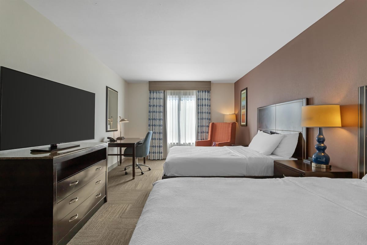 A hotel room with two beds, a desk, a television, and a window with curtains.