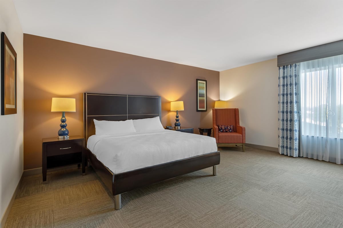 A hotel room with a large bed, two bedside tables with lamps, a chair, and a window with curtains.