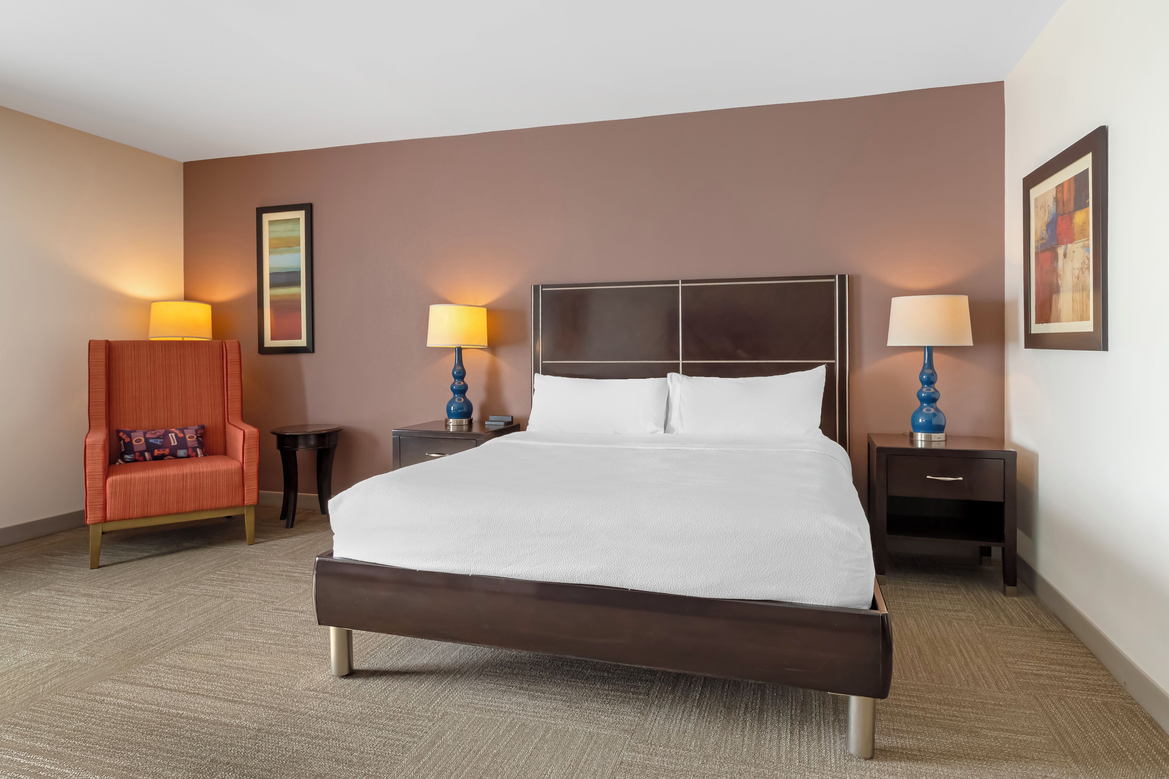 A hotel room with a large bed, two bedside tables with lamps, a chair, and a framed picture on the wall.