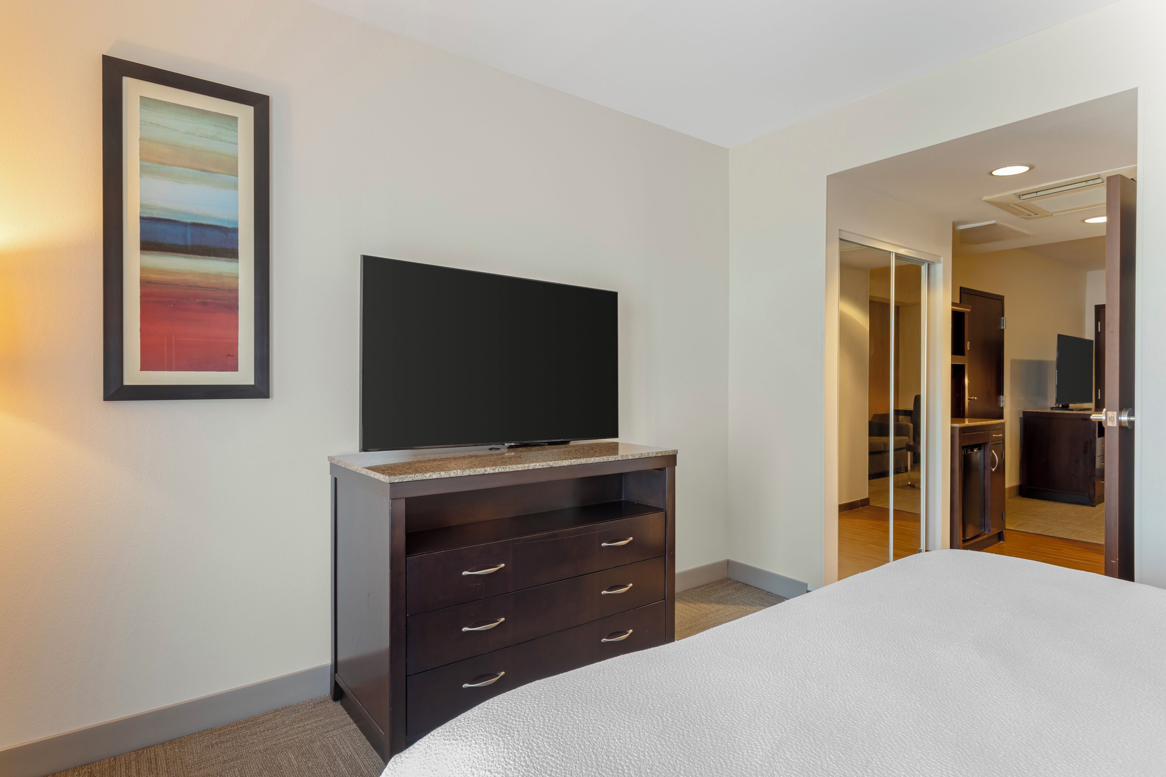 Une chambre d’hôtel avec un lit, une commode en bois foncé dotée de tiroirs, un téléviseur à écran plat fixé au mur et un tableau encadré.