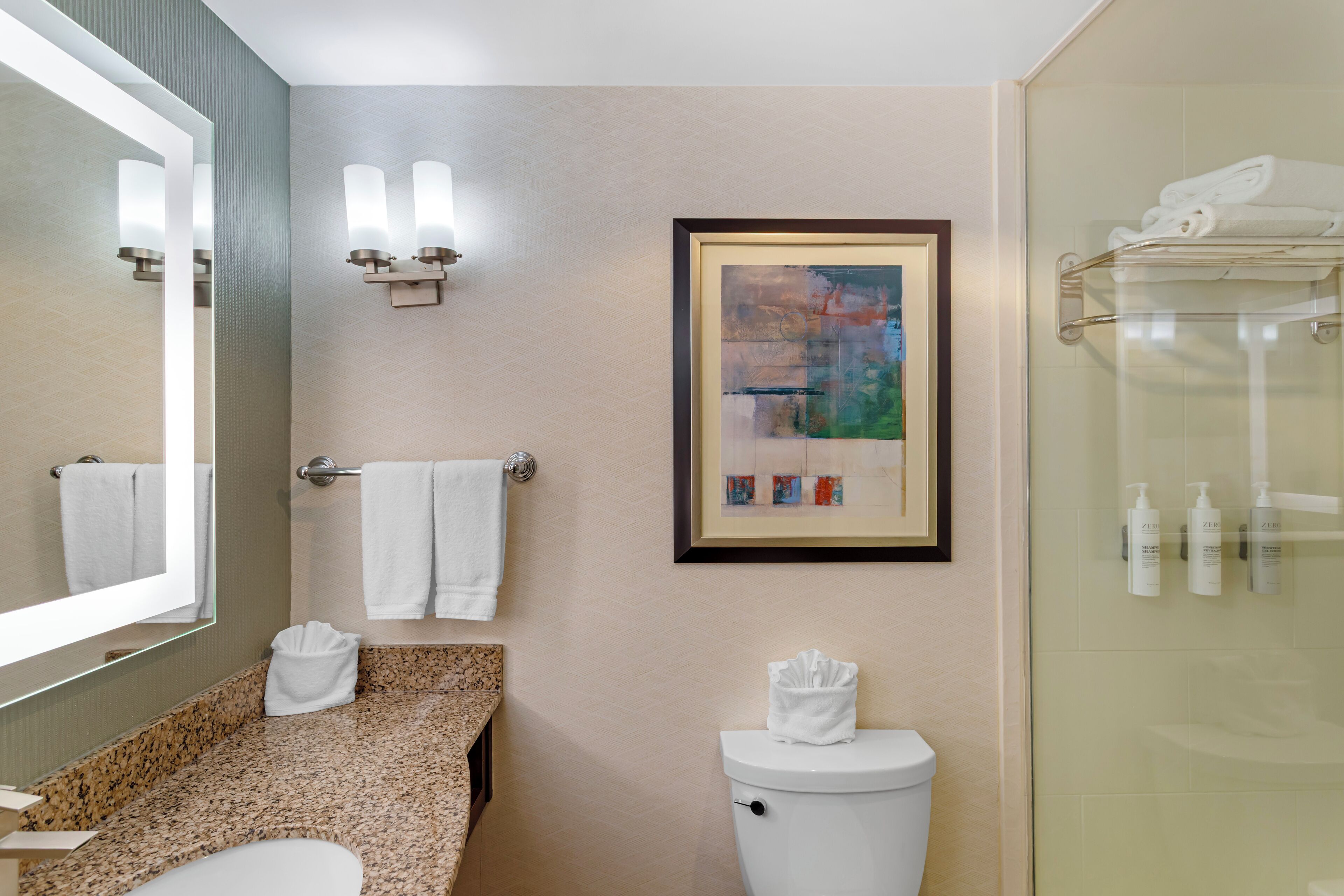 A bathroom with a granite countertop, a white toilet, a framed abstract painting, and a glass shower enclosure.