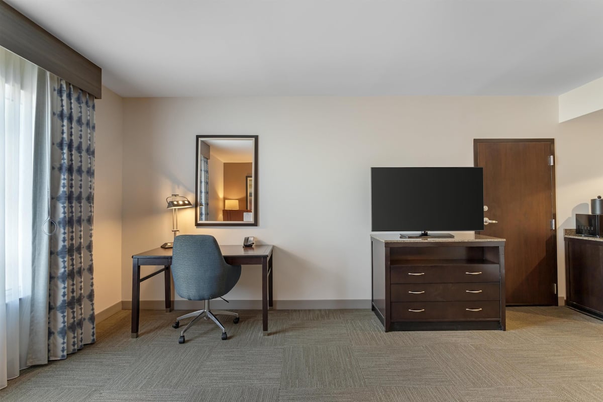 A hotel room with a desk, chair, television, wardrobe, and a mirror.