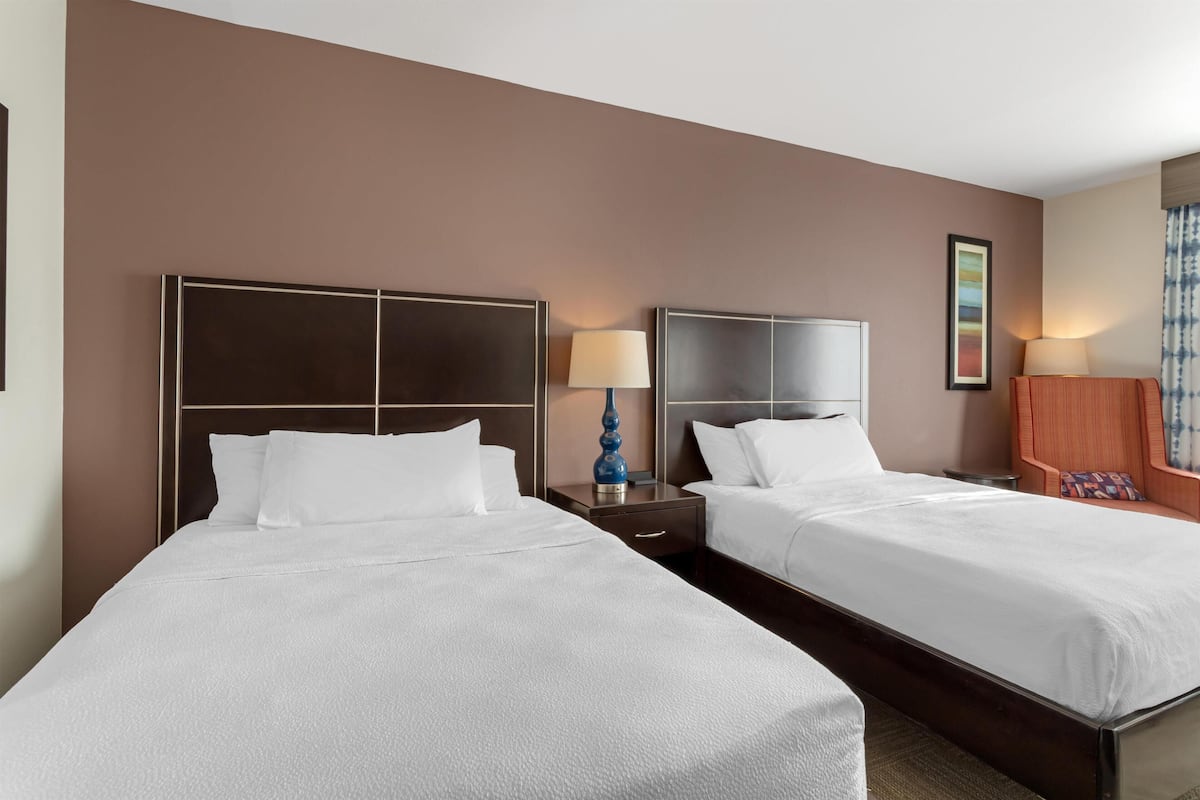 A hotel room with two beds, a nightstand with a lamp, a framed picture on the wall, and a window with curtains.