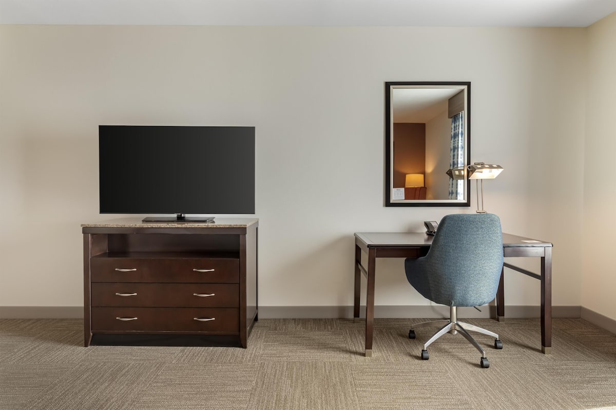 A hotel room with a flat-screen TV, a wooden desk, a blue office chair, a mirror, and a lamp.