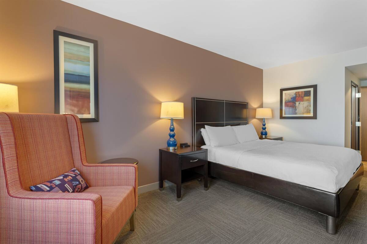 A hotel room with a bed, a chair, a nightstand with a lamp, a desk, and a wall-mounted TV.