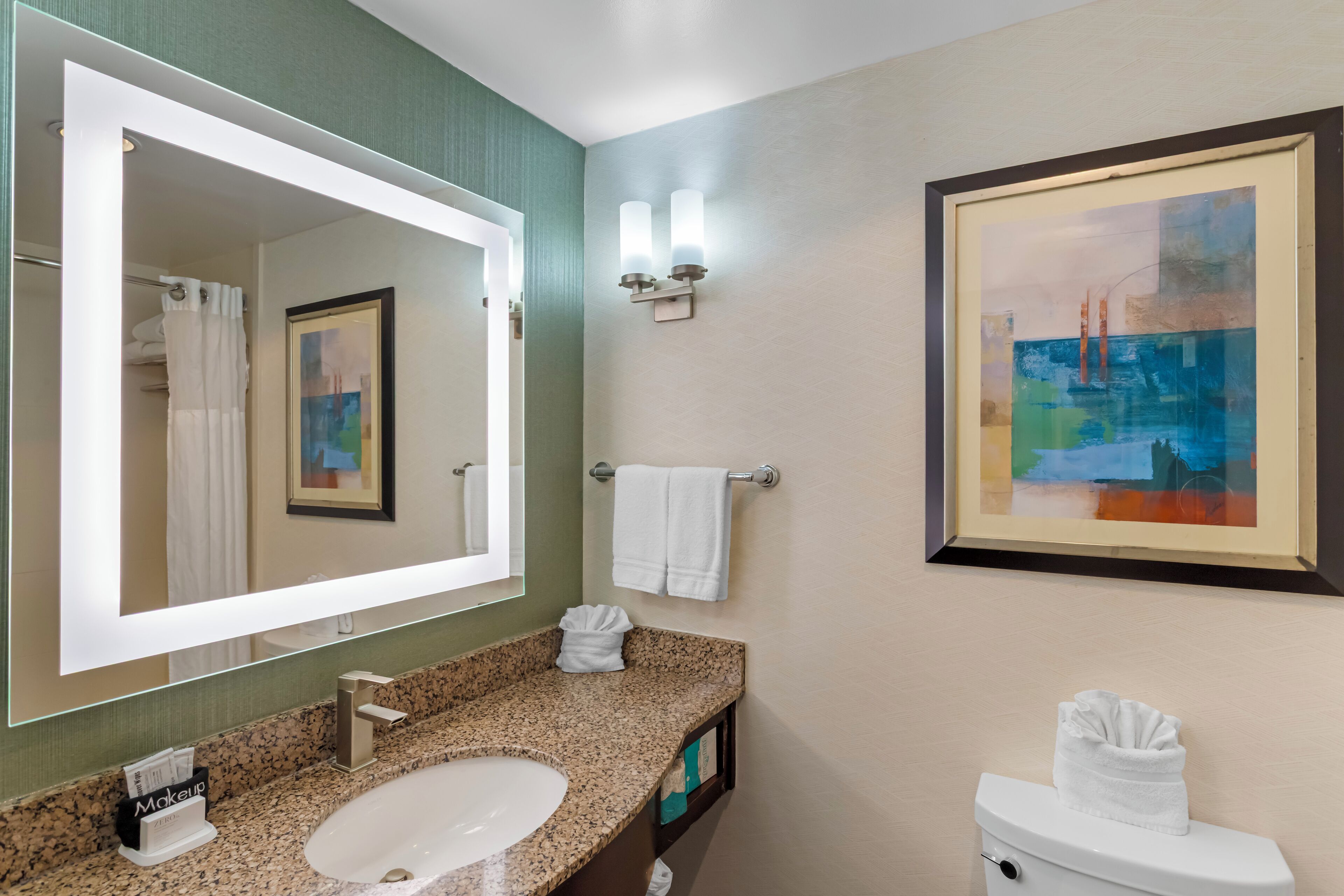 Une salle de bain avec un lavabo doté d’un plan de travail en granit, un grand miroir, un tableau encadré et un porte-serviettes.