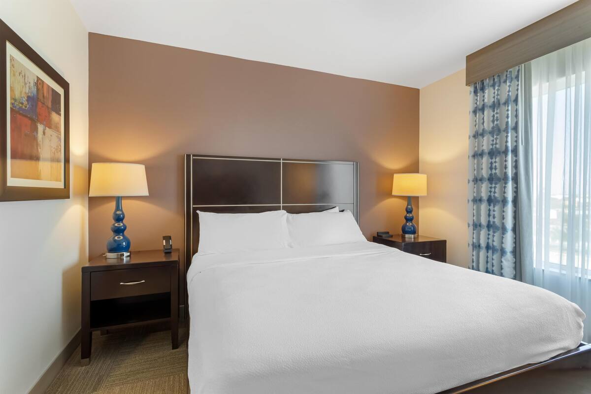 A hotel room with a large bed, two bedside tables with lamps, a headboard, and a window with curtains.