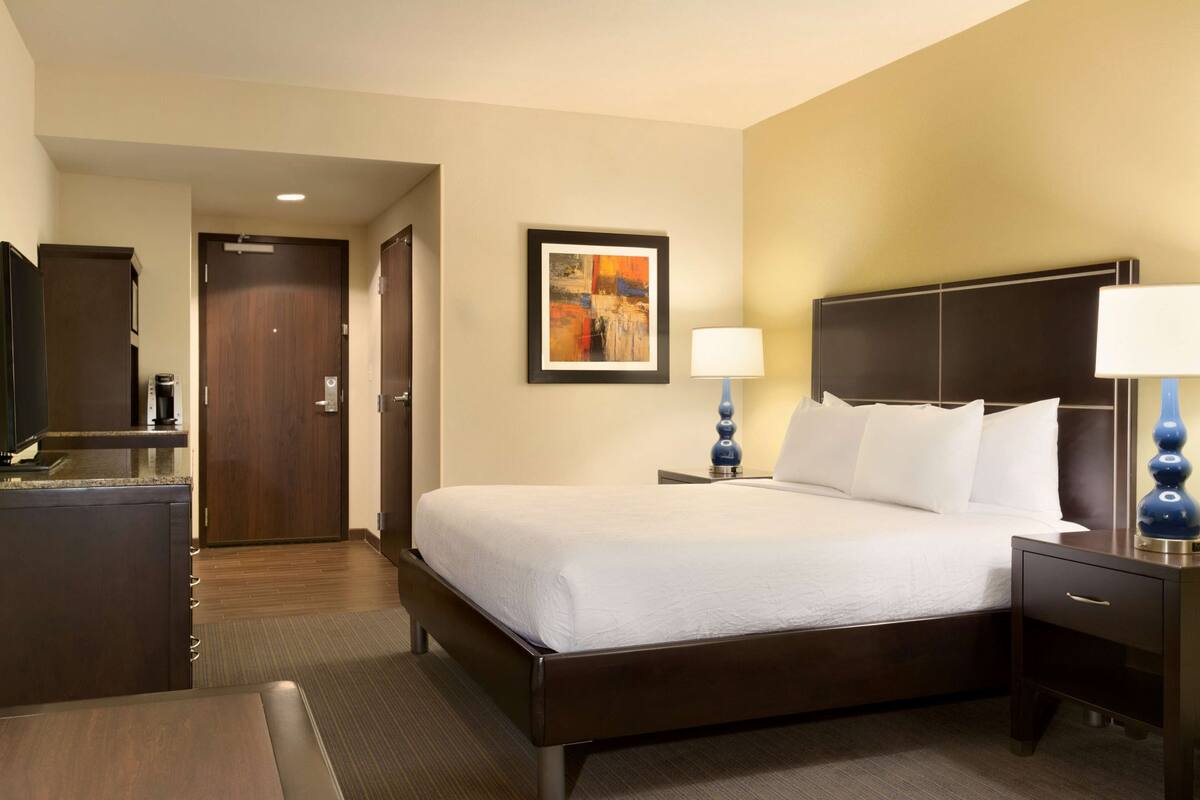 A hotel room with a bed, bedside tables, a desk, a chair, a framed artwork on the wall, and a door leading to another room.