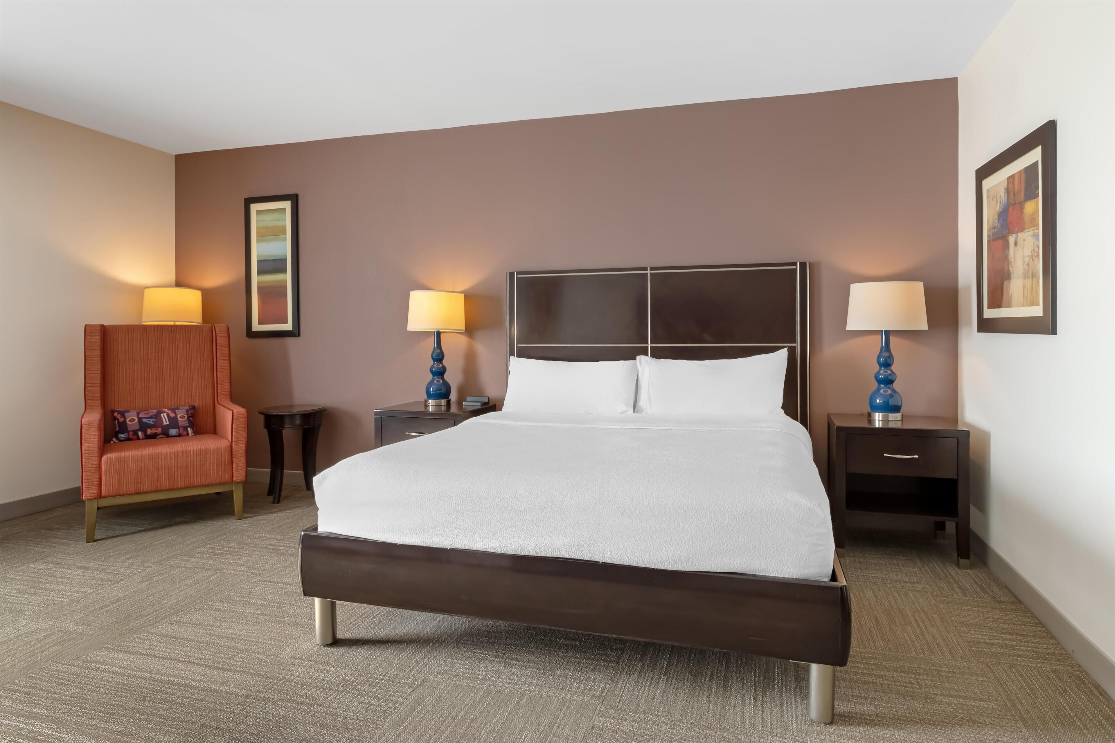 Habitación de hotel con una cama grande, dos mesitas de noche con lámparas, una silla y un cuadro en la pared.
